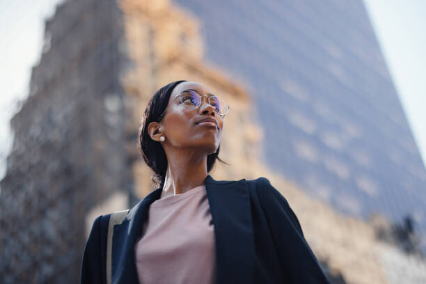 Confident businesswoman wearing glasses, standing in a bustling city with tall buildings in the background, representing success and ambition.