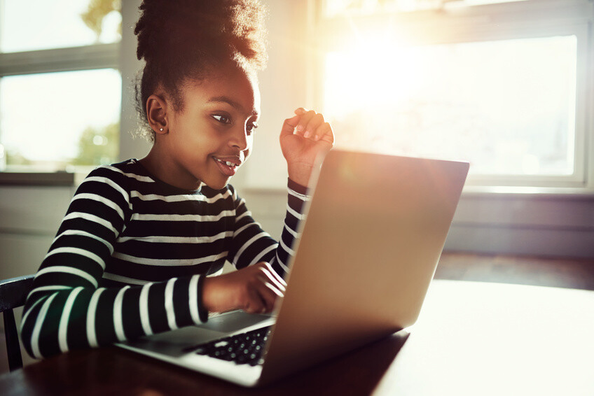 little ethnic girl sitting at home at a table working on her homework on a laptop computer reading the screen with a thoughtful expression